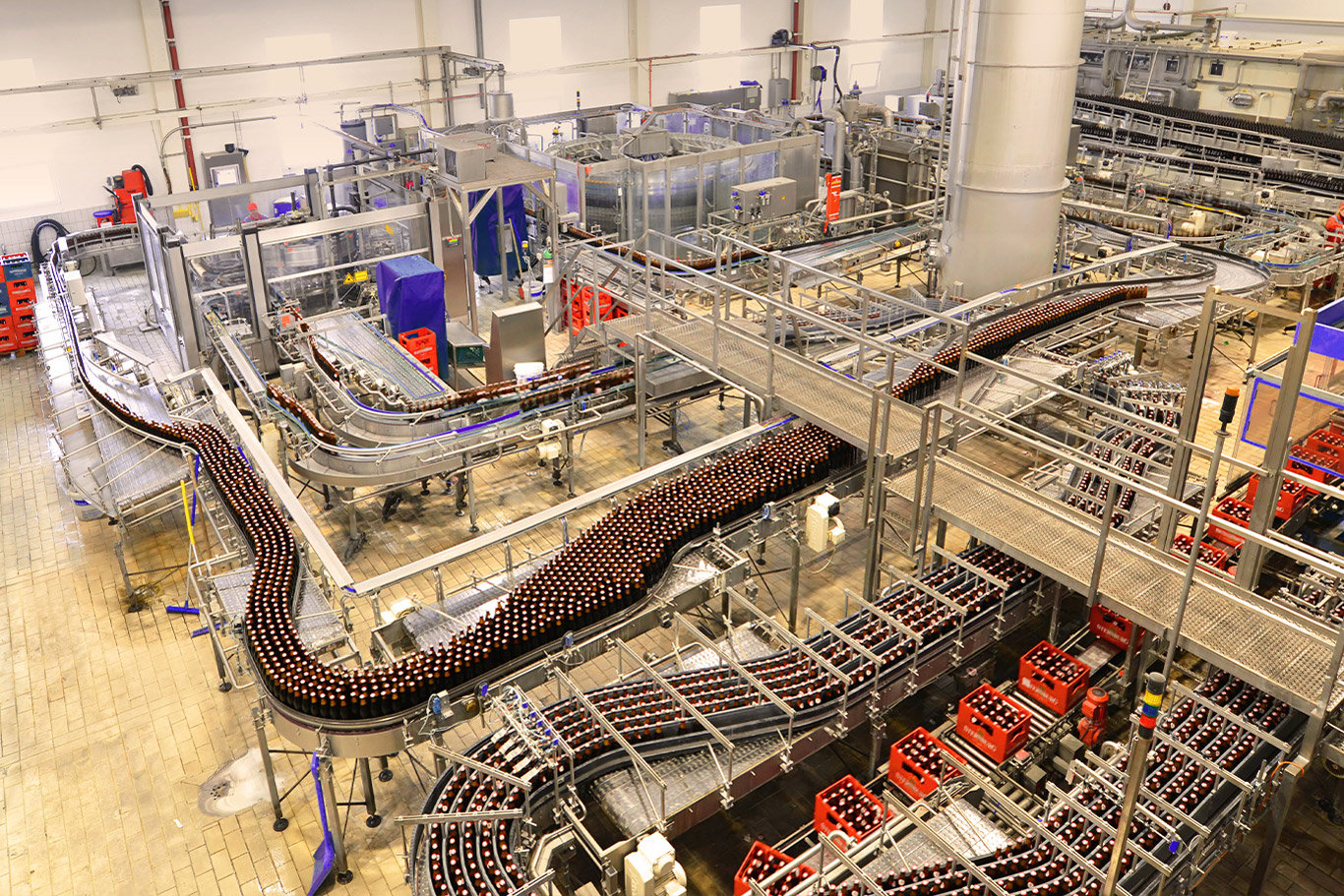 Wide-angle view of a modern beverage manufacturing plant with automated bottling lines, stainless steel processing equipment, and high-speed conveyors transporting bottles through production.